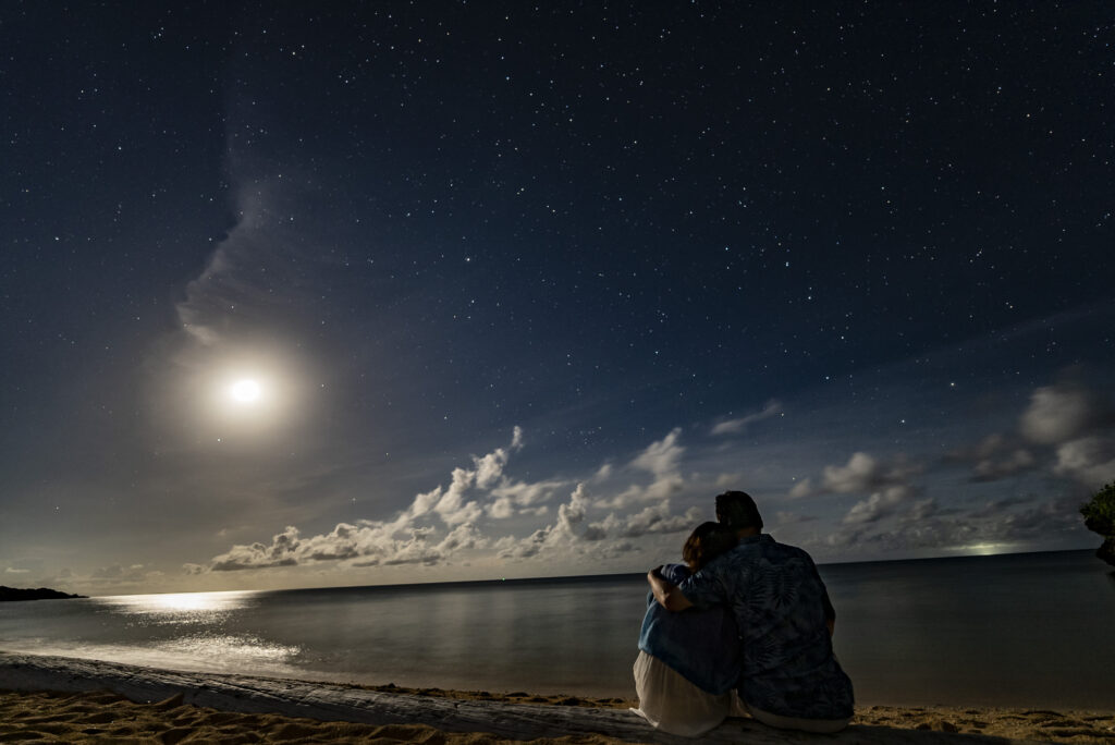 Couple sitting on beach watching moonrise over the ocean under starry sky at Ishigaki Island, near Nagareboshi no Oka
